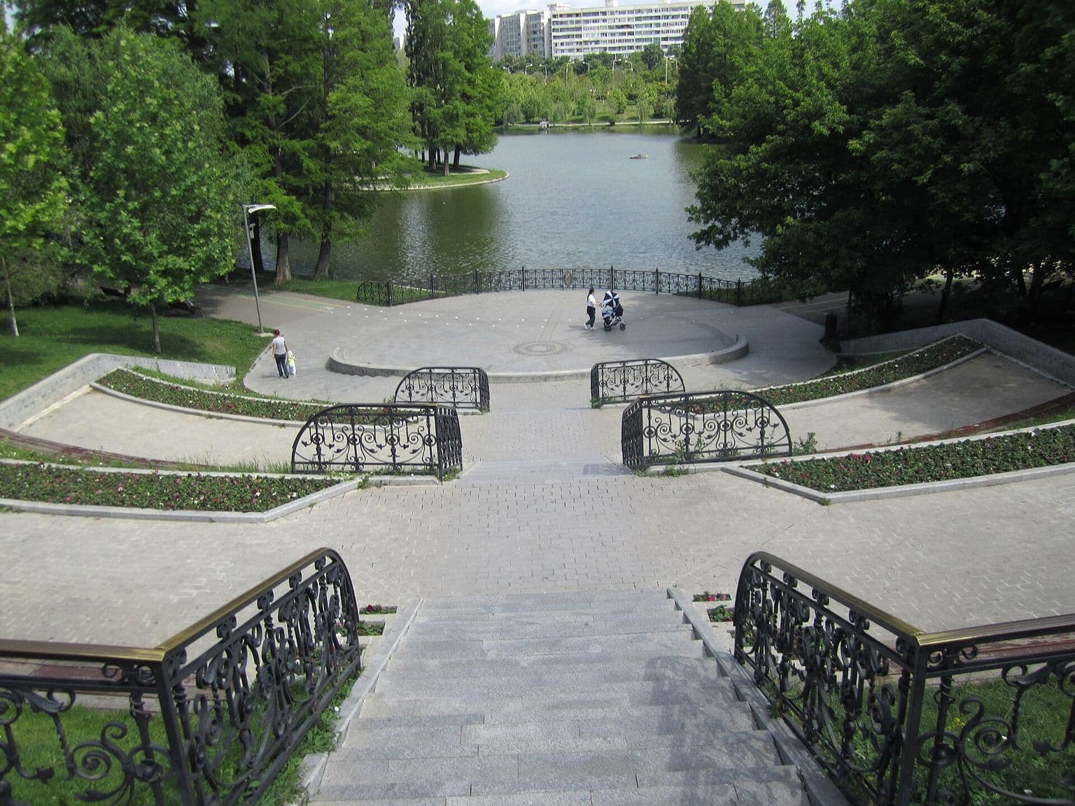 Scări largi de piatră coboară spre Lacul Titan din Parcul IOR, între copaci înfrunziți, cu blocuri în depărtare