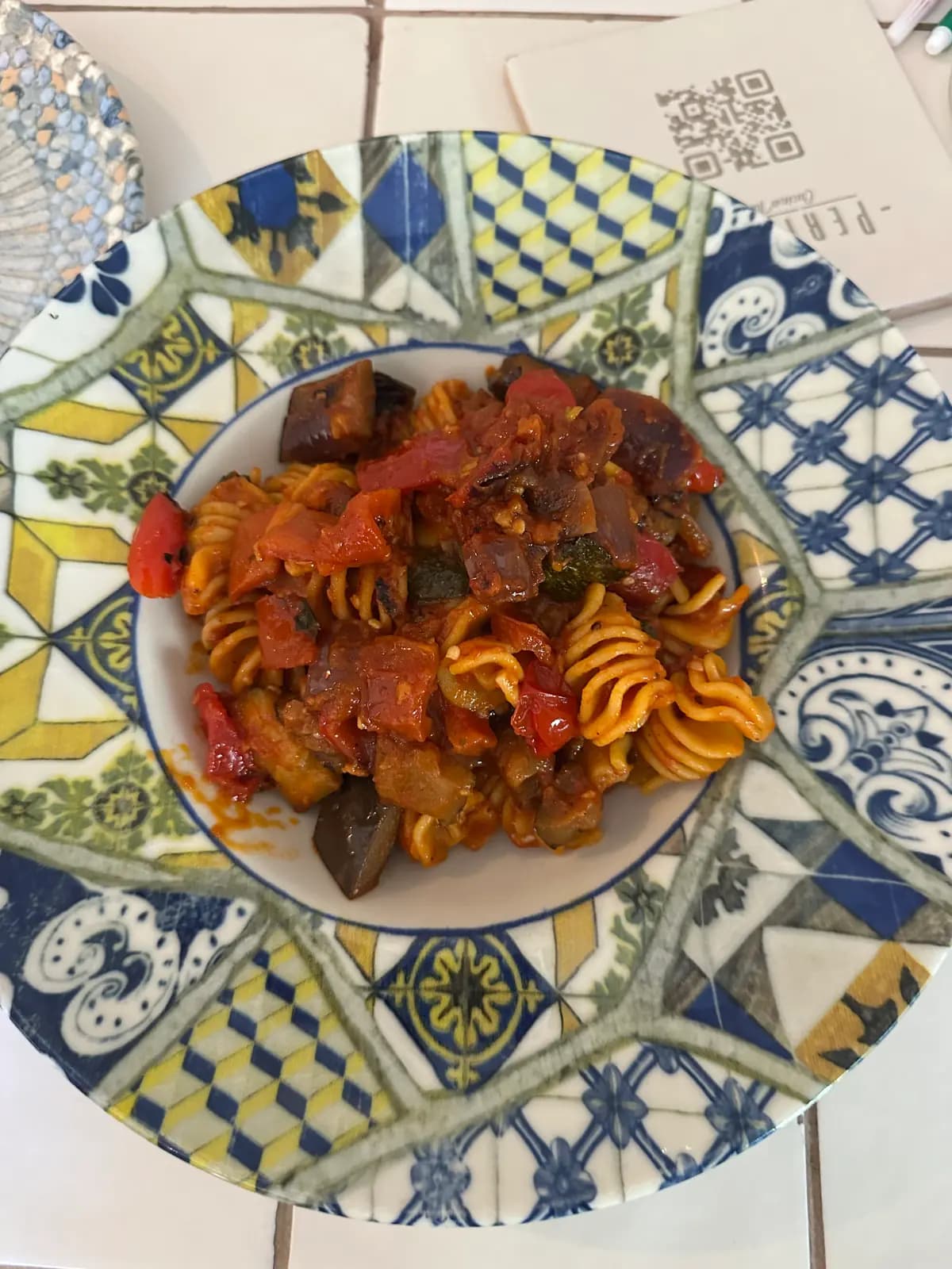 Des pâtes radiatori aux aubergines, courgettes et poivrons rôtis dans une sauce tomate, servies dans une assiette creuse aux motifs de carreaux bleus et jaunes, sur une table carrelée de blanc