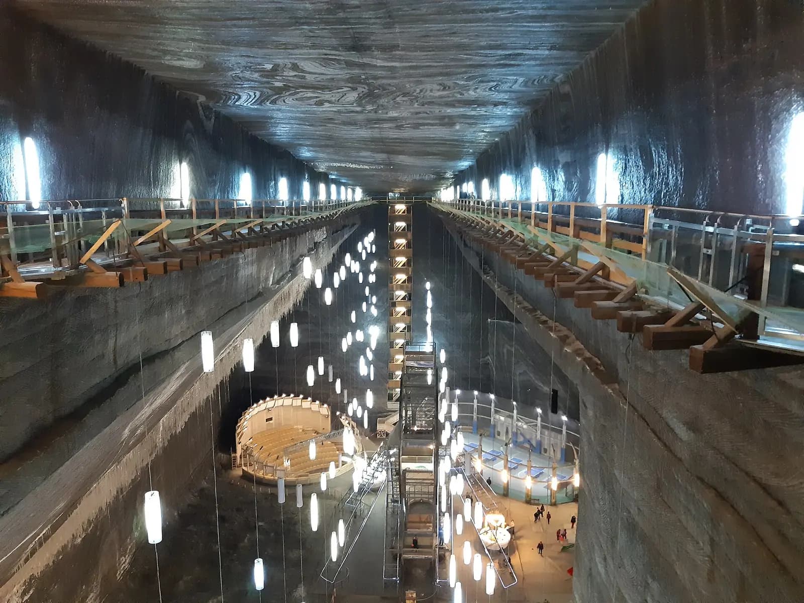 Mina Rudolf de la Salina Turda văzută de sus: pereți înalți de sare, galerii de lemn, șiruri de lămpi atârnate și, jos, amfiteatrul rotund, terenurile de joacă și turnul liftului
