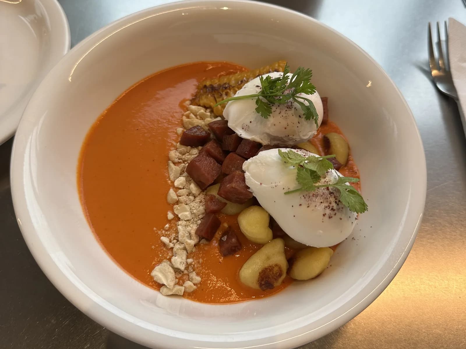 Bol la Eggcetera: două ouă poșate cu coriandru și sumac, cuburi de chorizo, gnocchi rumeniți, porumb copt și brânză sfărâmată, într-un sos cremos de ardei roșii, pe o masă de inox