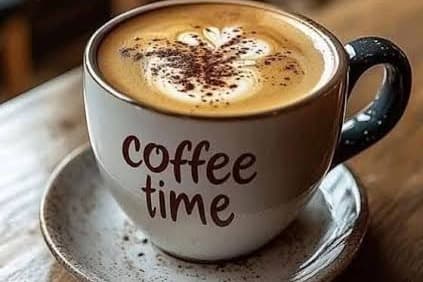 A white cup of cappuccino with a foam heart and a dusting of cocoa, "coffee time" written on it, on a saucer on a wooden table